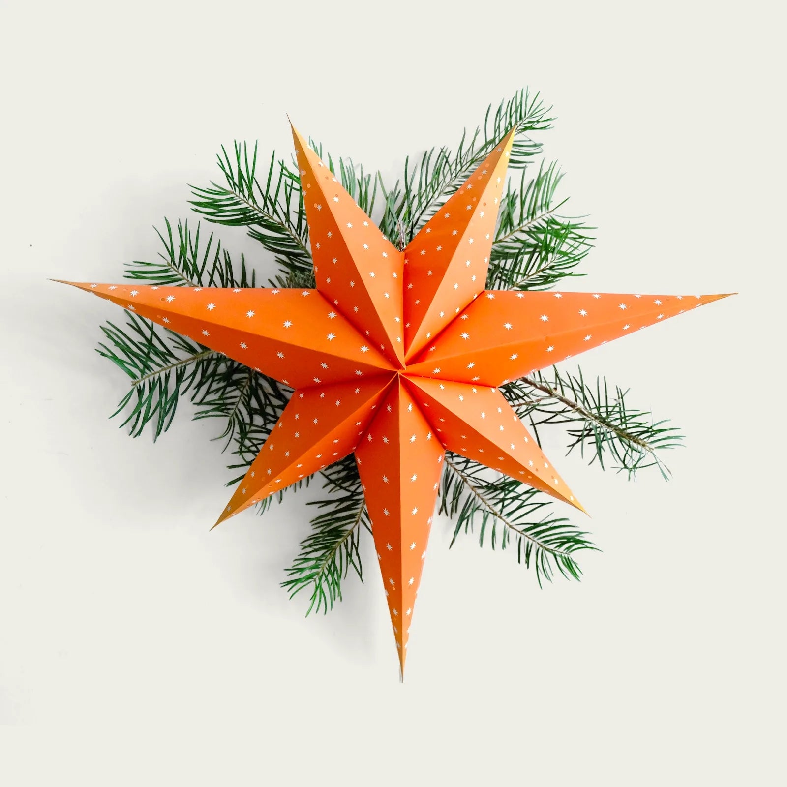 en-orange-adventssjarna-monstrad-med-vita-sma-stjarnor-i-bakgrunden-ligger-granris-som-dekoration